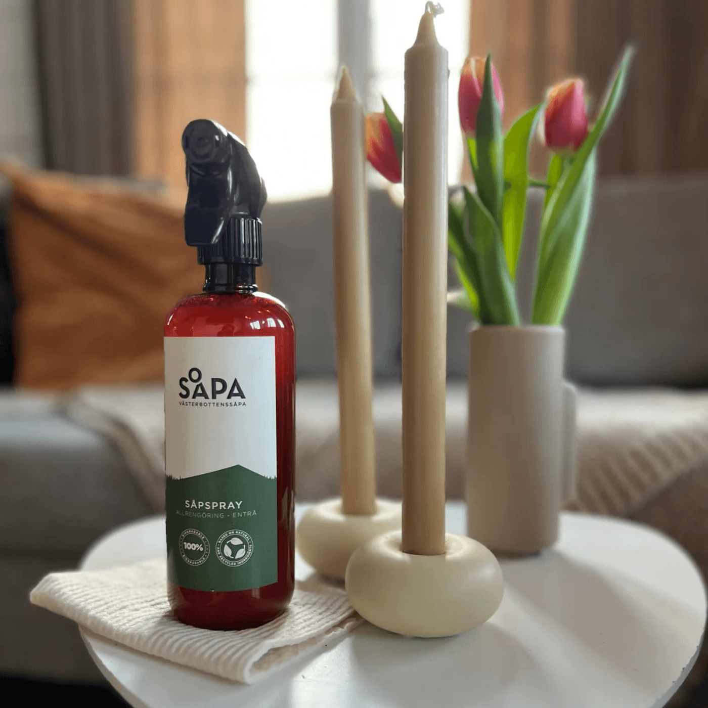 En flaska naturlig såpspray och en trasa placerade på ett bord i vardagsrummet med ljus och en vas med blommor.