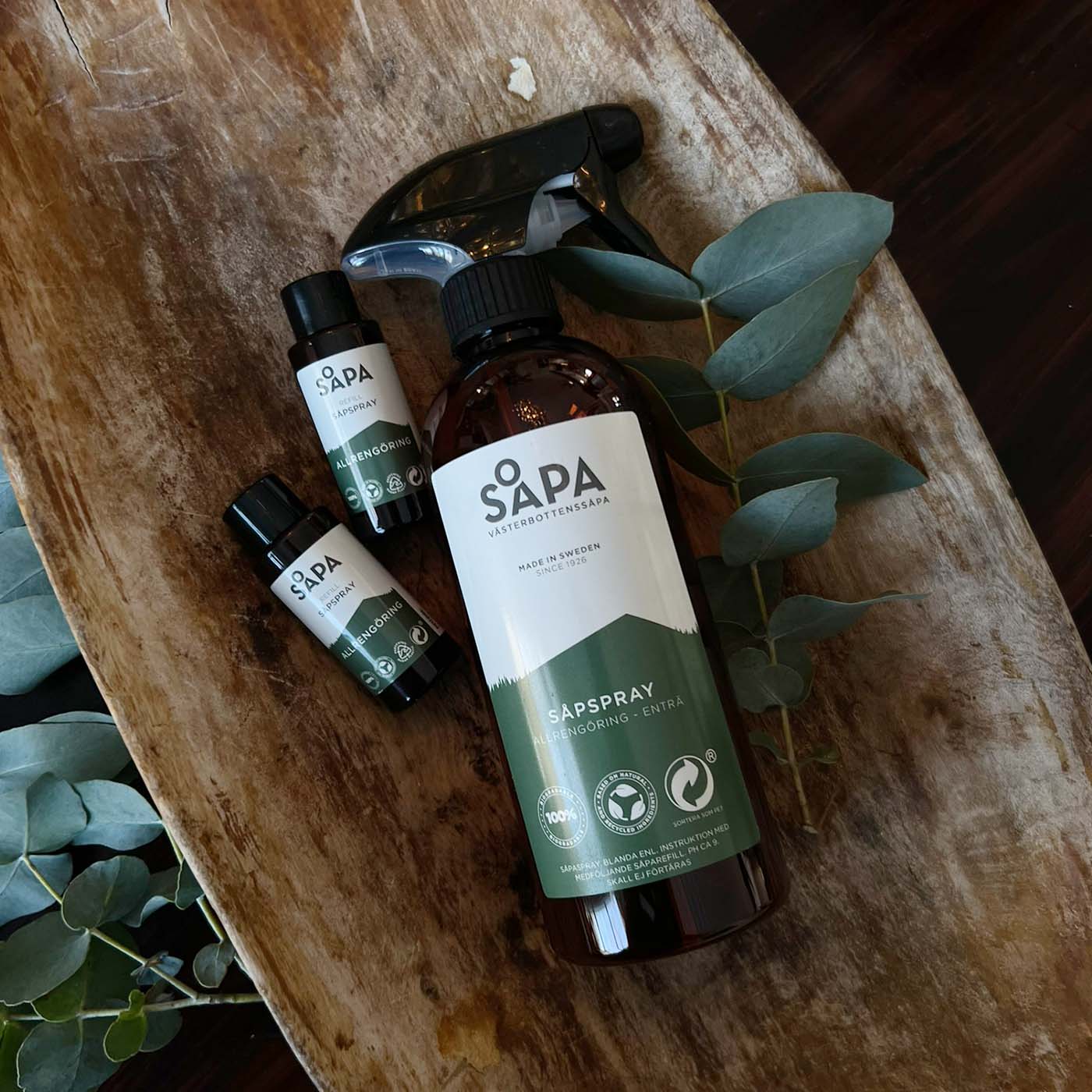 Flaska med Västerbottenssåpa Allrengöring Enträ med två refiller liggande på en rustik träyta. Naturlig, effektiv och skonsam rengöring.