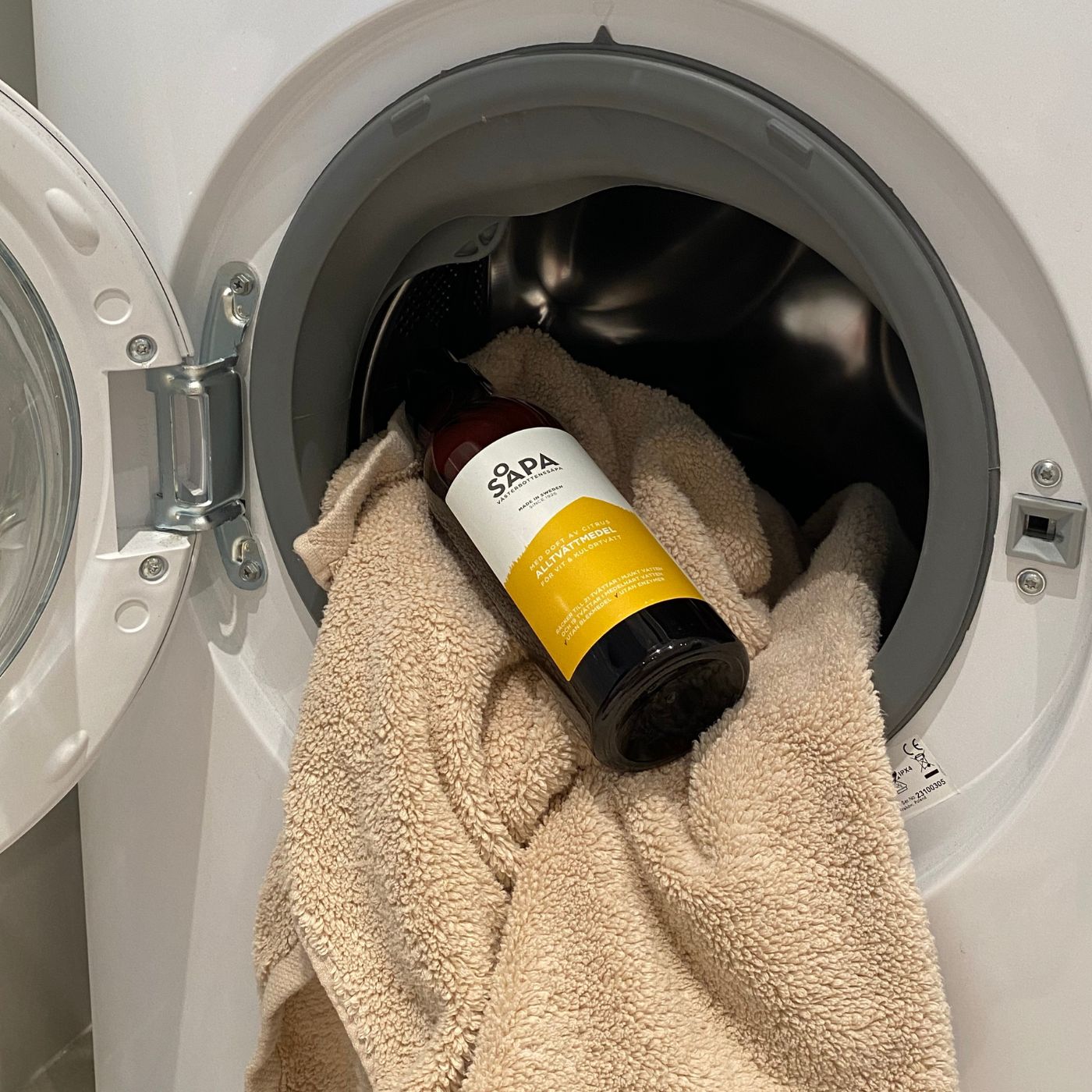 Flaska med citrusdoftande tvättmedel placerad i en tvättmaskin tillsammans med en beige handduk. Miljövänligt och skonsamt för vit- och kulörtvätt