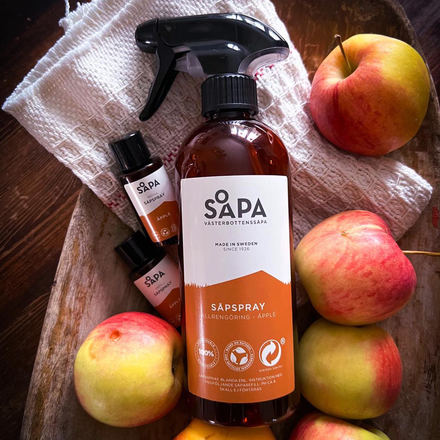 Flaska med Västerbottenssåpa äpple såpspray och två refiller placerad i en rustik trälåda tillsammans med röda äpplen. 