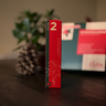 Rött och grönt paket märkt med siffran 2 för andra advent ur Västerbottenssåpas adventskalender, med en kotte och presentask i bakgrunden.