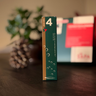 Grönt paket märkt med siffran 4 för fjärde advent ur Västerbottenssåpas adventskalender. En kotte och kalenderns ytterförpackning syns i bakgrunden.