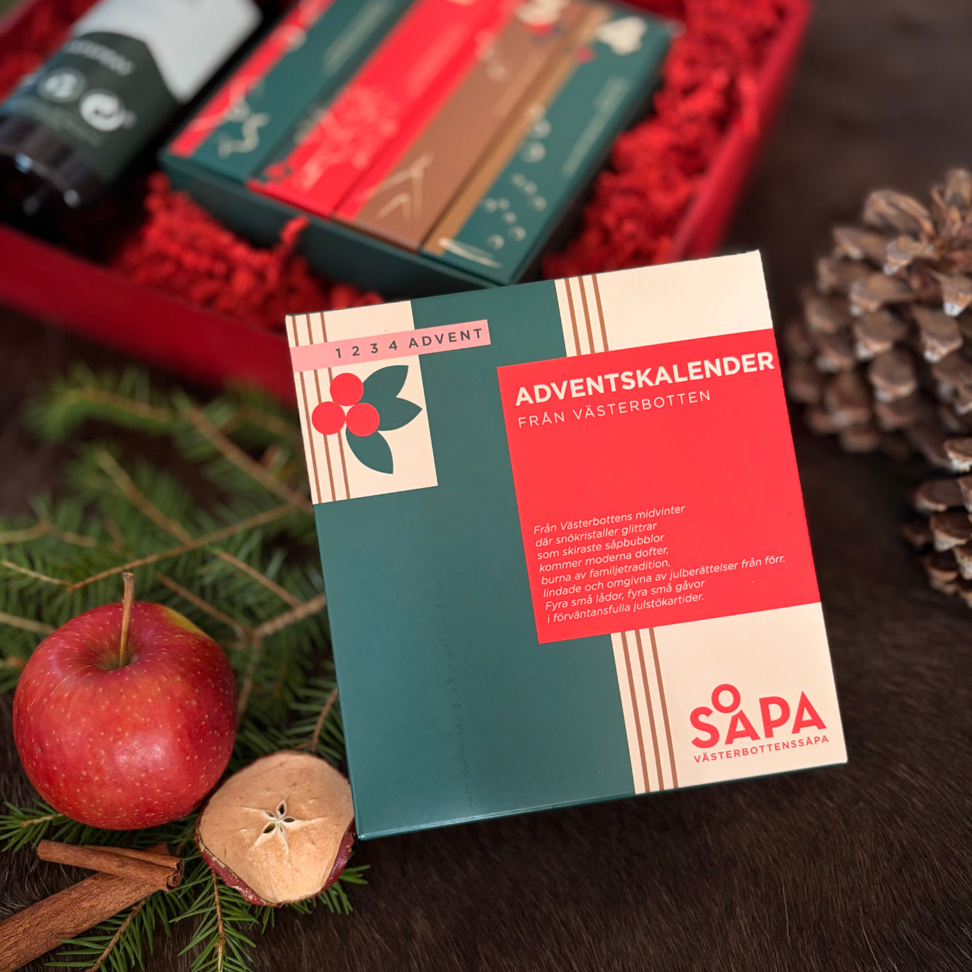 Västerbottenssåpas gröna adventskalender med texten ”1 2 3 4 advent” ligger bredvid granris, äpplen, kanelstänger och en kotte. I bakgrunden syns de fyra adventspaketen.