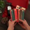 Händer håller fyra numrerade adventspaket från Västerbottenssåpas adventskalender, med en öppen röd presentask och sprayflaska i bakgrunden.