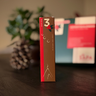 Brunt och rött paket märkt med siffran 3 för tredje advent ur Västerbottenssåpas adventskalender, med en kotte och presentask i bakgrunden.